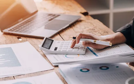 Manos usando una calculadora sobre papeles con gráficos circulares, junto a una notebook
