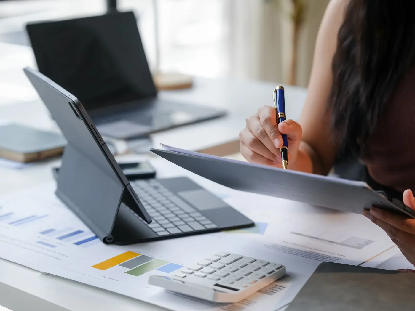Una mujer con una lapicera revisa una carpeta frente a una tablet, una calculadora y planillas con gráficos