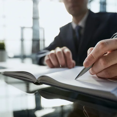 Un hombre de traje escribe en un cuaderno abierto sobre un escritorio vidriado