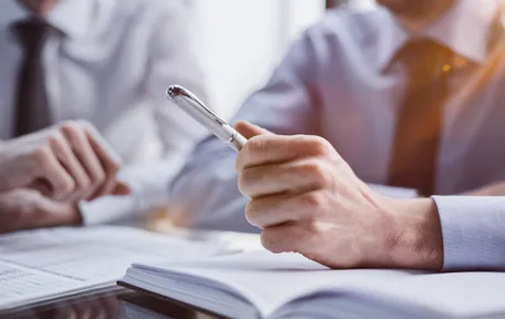 Dos personas de camisa y corbata conversan con una lapicera y documentos sobre la mesa