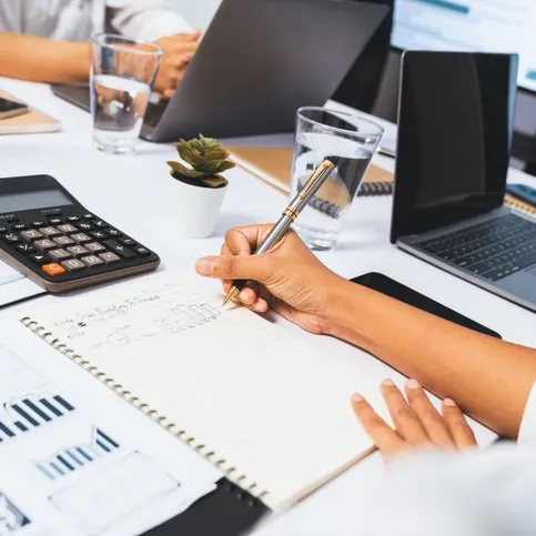 Una persona anota en un cuaderno en una mesa de reunión con notebooks, una calculadora y planillas