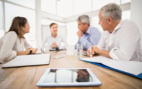 Cuatro personas conversan alrededor de una mesa de reunión, con una tablet en primer plano
